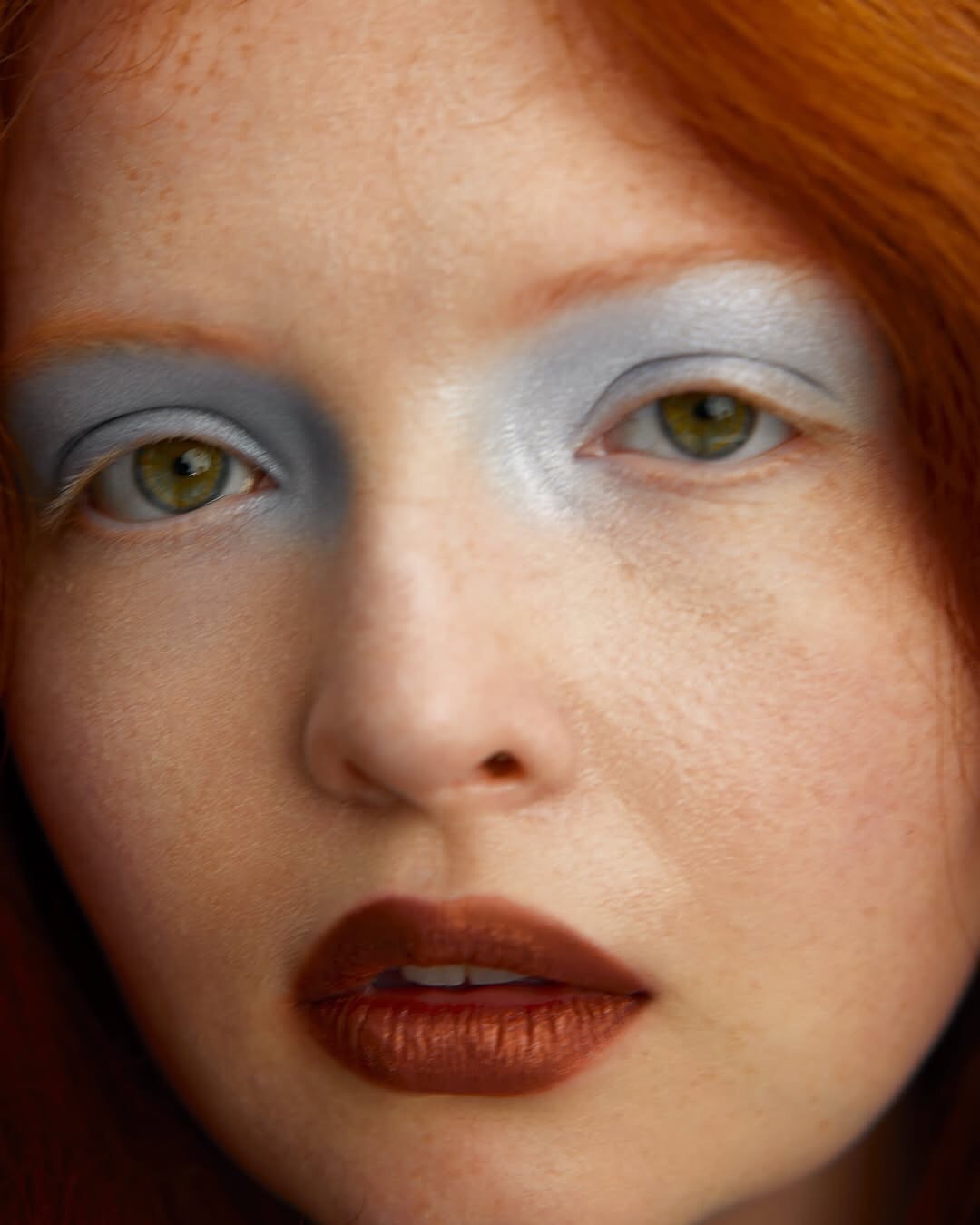 Close-up portrait of a red-haired adult female with green eyes wearing light blue eyeshadow and dark red lipstick in a neutral indoor setting.