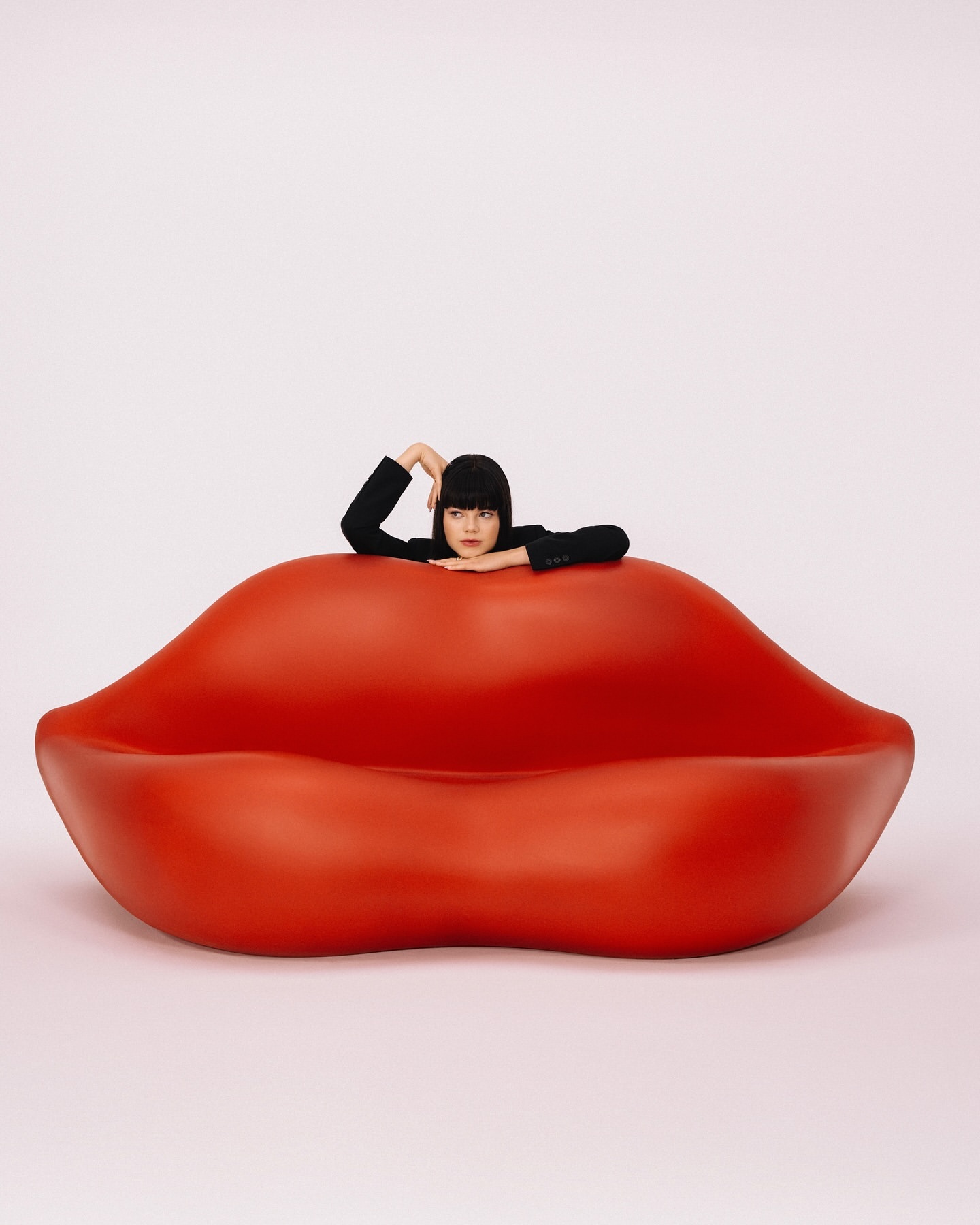 Woman in a black blazer lounges in a bright red, oversized lips couch against a neutral backdrop in a minimalist, studio portrait.