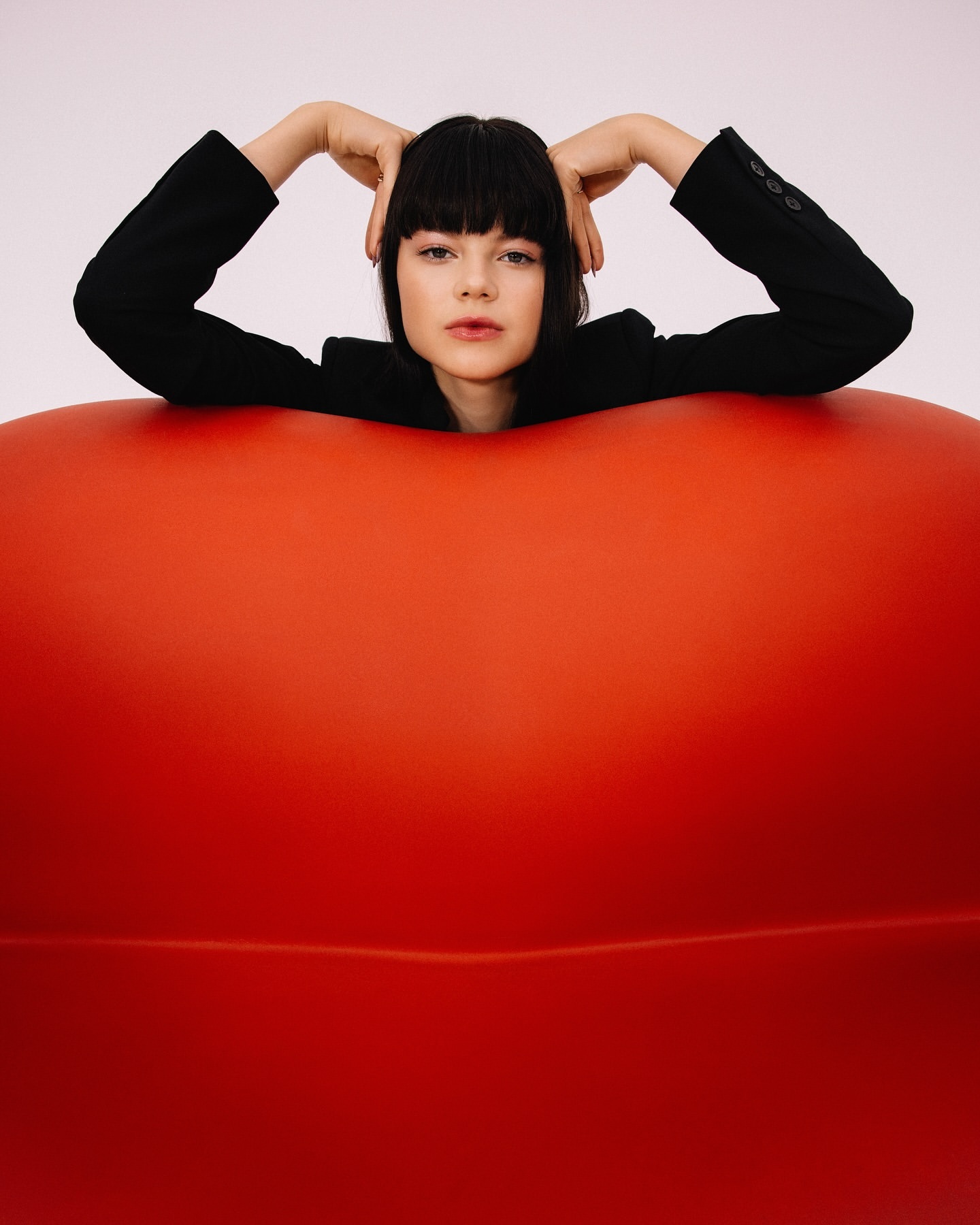 A young woman with dark hair and a black blazer poses on a red, rounded couch against a white background in a studio setting.