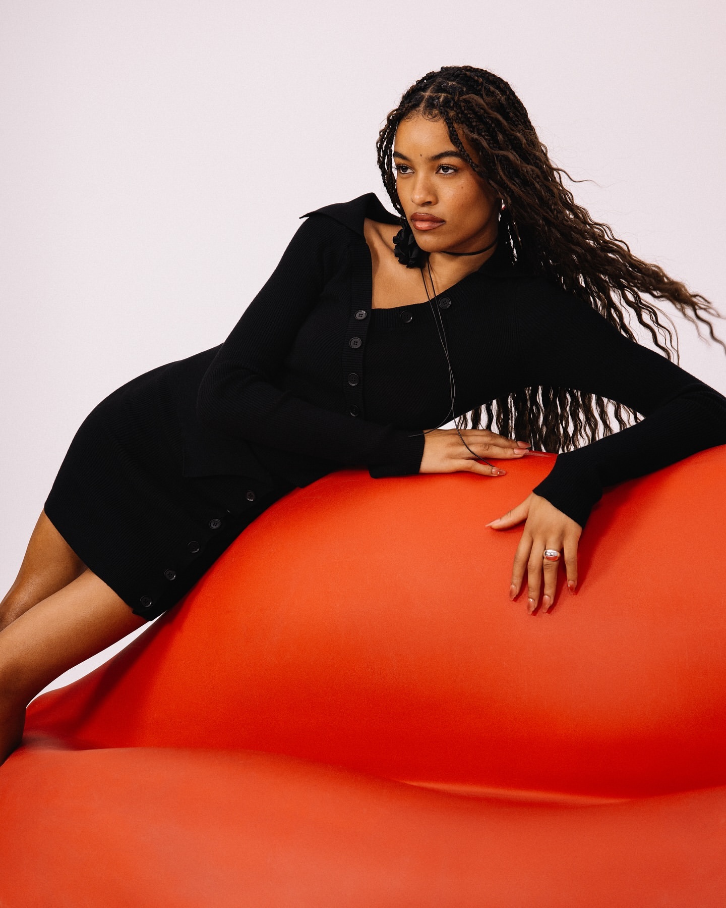 Woman in a black long-sleeved dress poses against an orange backdrop in a studio. Her long, wavy hair is parted in the middle.