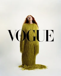 Model with long red hair wearing a chartreuse fringe dress standing against a plain white seamless background