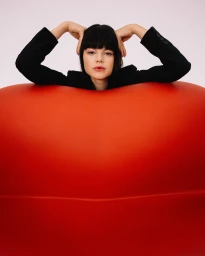 A woman with dark hair and bangs resting her hands behind her head, leaning over a smooth, vibrant red curved structure against a plain white background.