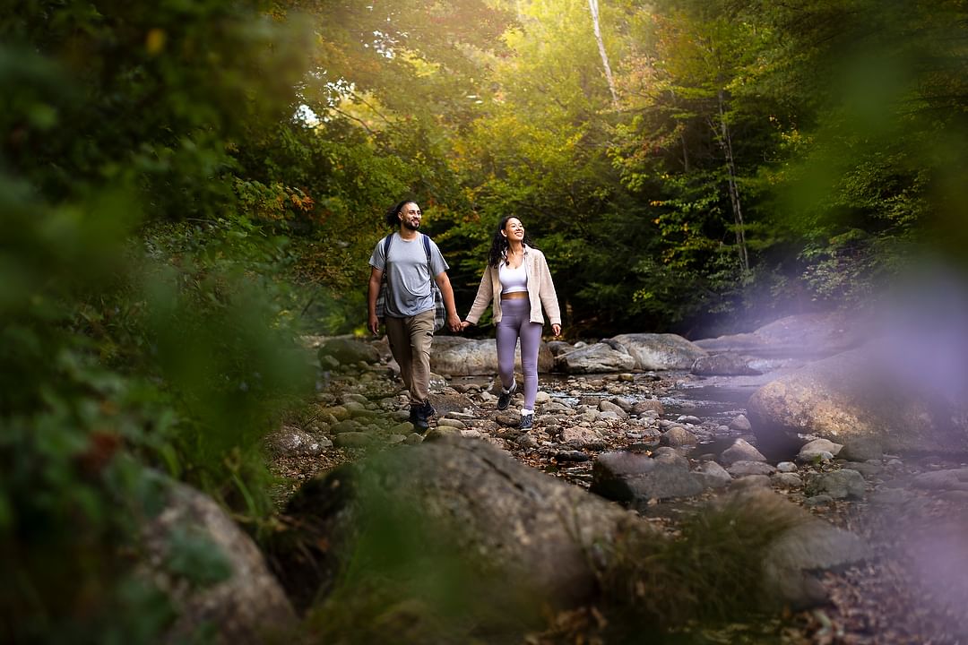 A couple hikes hand-in-hand on a rocky stream bed in a forest. The sun shines through the trees creating a peaceful outdoor scene.