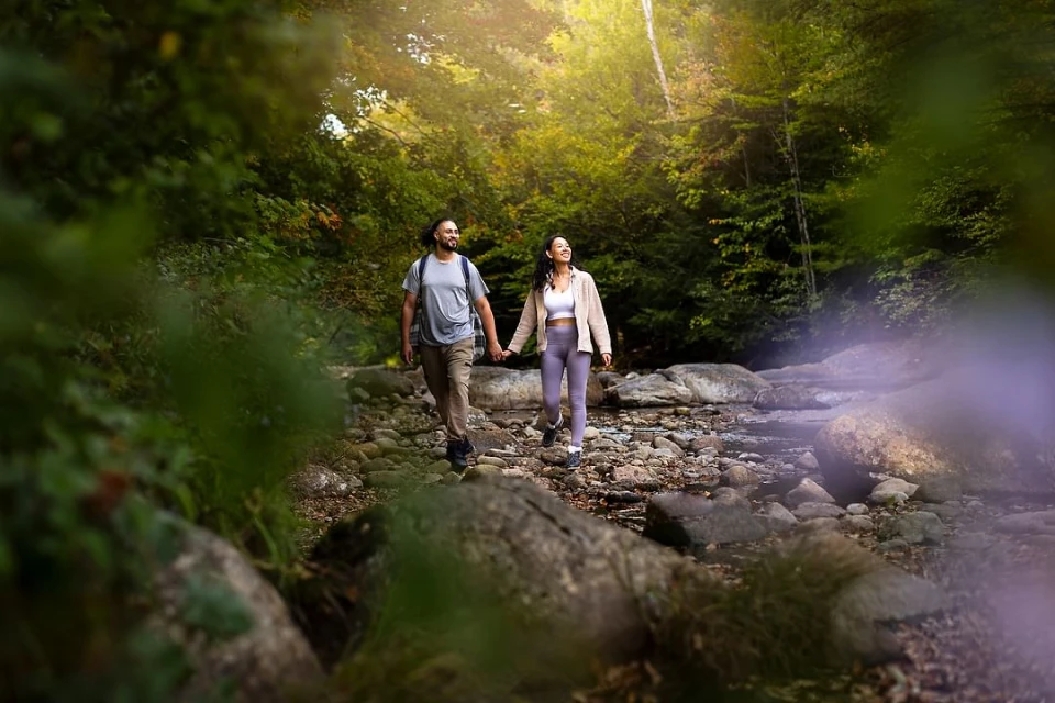 A couple hikes hand-in-hand on a rocky stream bed in a forest. The sun shines through the trees creating a peaceful outdoor scene.