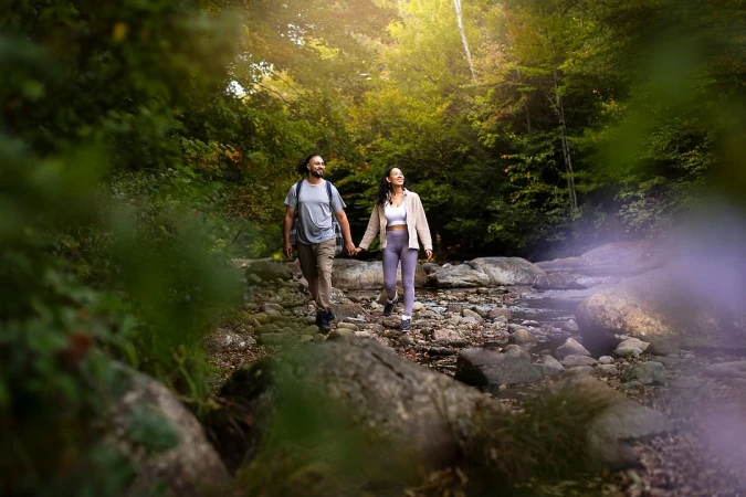 A couple hikes hand-in-hand on a rocky stream bed in a forest. The sun shines through the trees creating a peaceful outdoor scene.