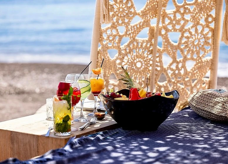Beachside table with cocktails and fruit bowl on a blue patterned cloth, set against a backdrop of sand, sea, and crocheted canopy.