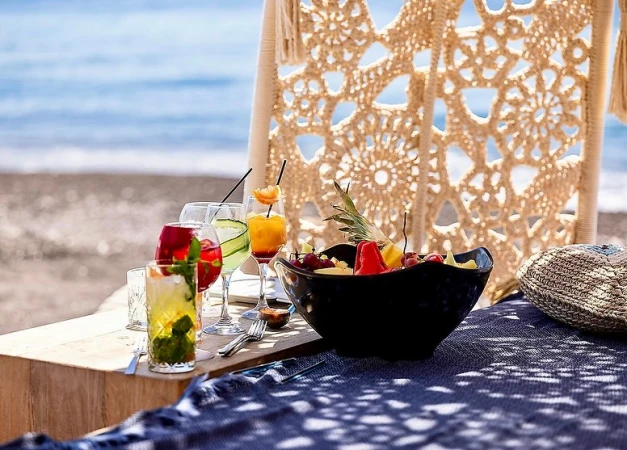 Beachside table with cocktails and fruit bowl on a blue patterned cloth, set against a backdrop of sand, sea, and crocheted canopy.