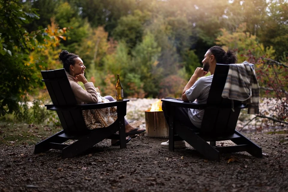 Couple relaxing by the campfire: A woman and a man sit in Adirondack chairs, enjoying a drink and each other's company in nature.