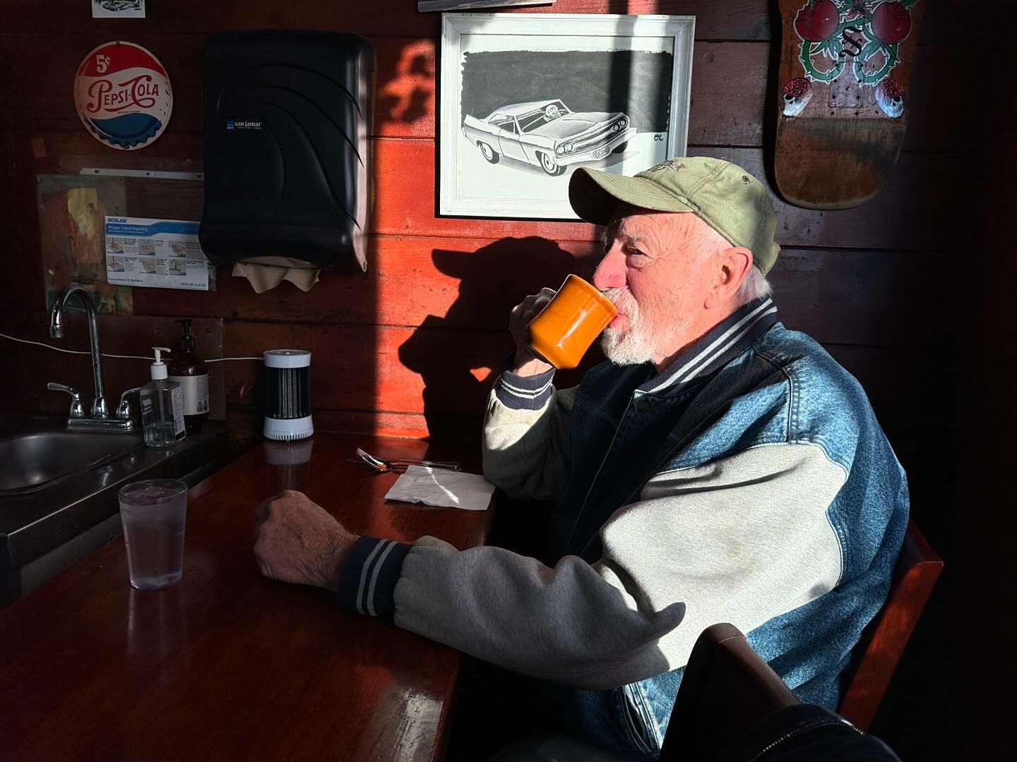 Elderly man in cap sips from mug at wooden table in warm light, with vintage signs, sink, and artwork in the background, indoors.
