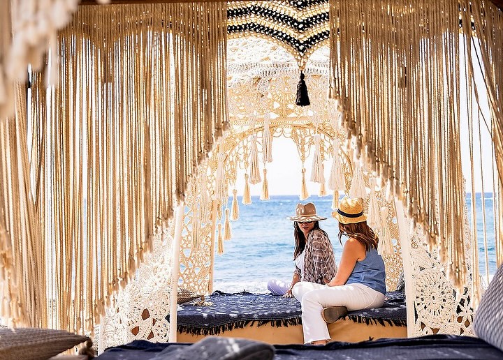 Two women in hats sit near the ocean in an elaborate macrame structure, enjoying the seaside view with tan sand and blue water.