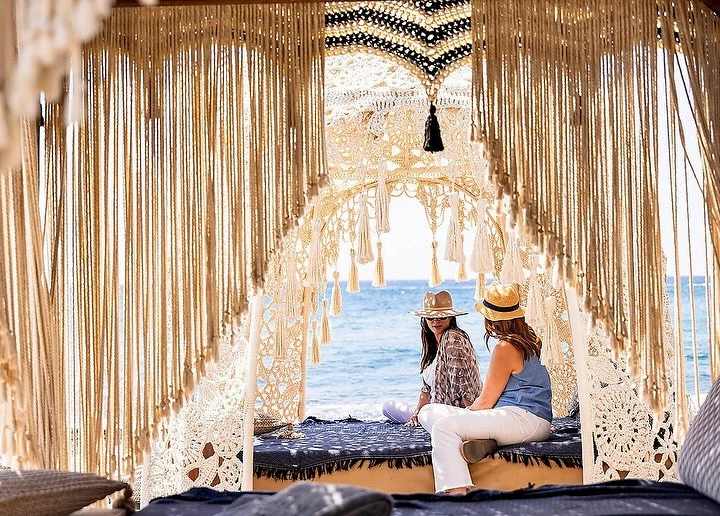 Two women in hats sit near the ocean in an elaborate macrame structure, enjoying the seaside view with tan sand and blue water.