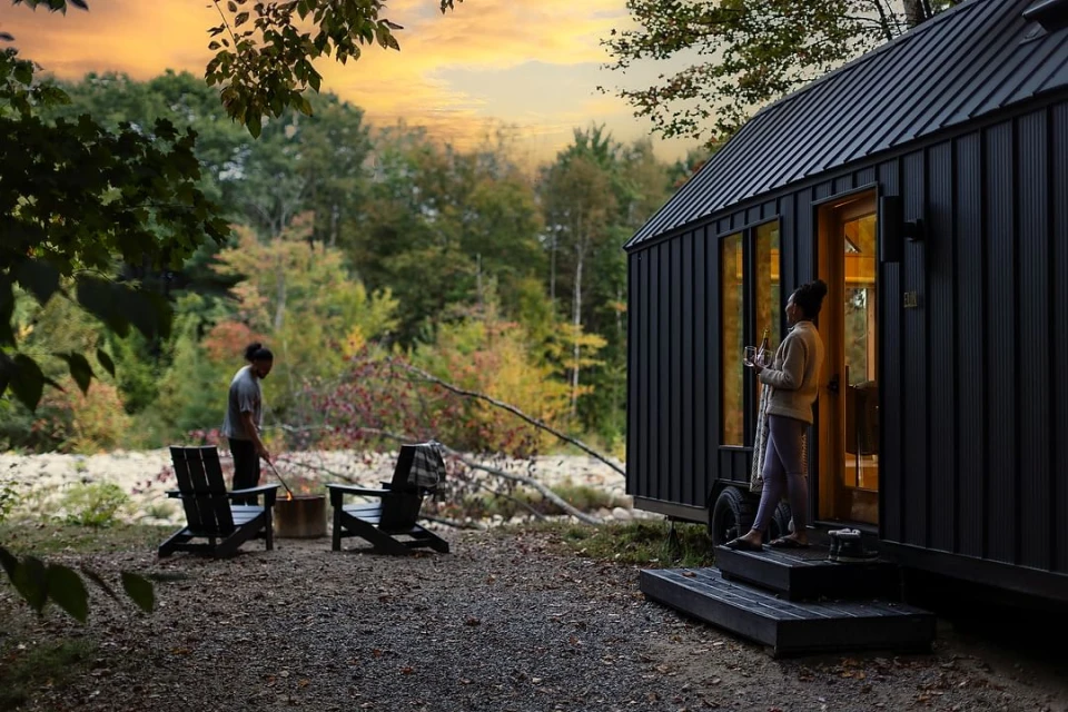 Two people at a tiny home in a nature setting with autumn colors during sunset. One woman on the porch, one man near a fire pit.