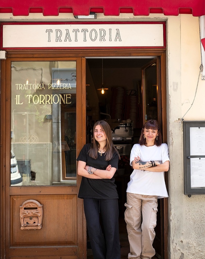 Two women stand in the doorway of a trattoria, smiling with arms crossed. The sign reads "Trattoria Pizzeria Il Torrione".