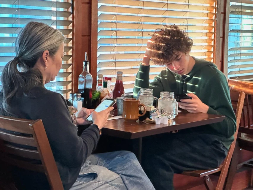 Adult woman and teenage male use smartphones at a wooden table inside a restaurant with slatted blinds and visible condiments and drinks.