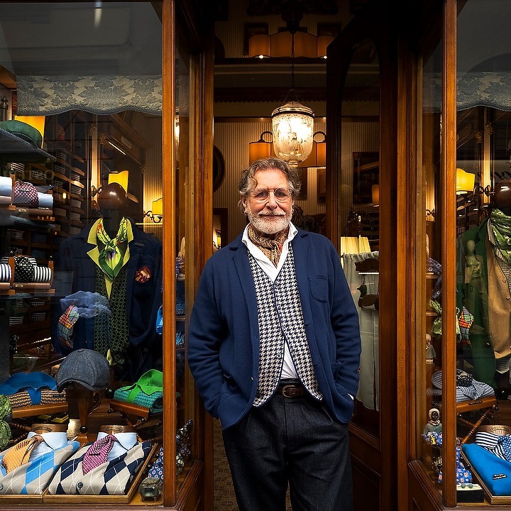 Portrait of an older gentleman in a blue blazer, posing in the doorway of a bespoke clothing shop, surrounded by display cases.