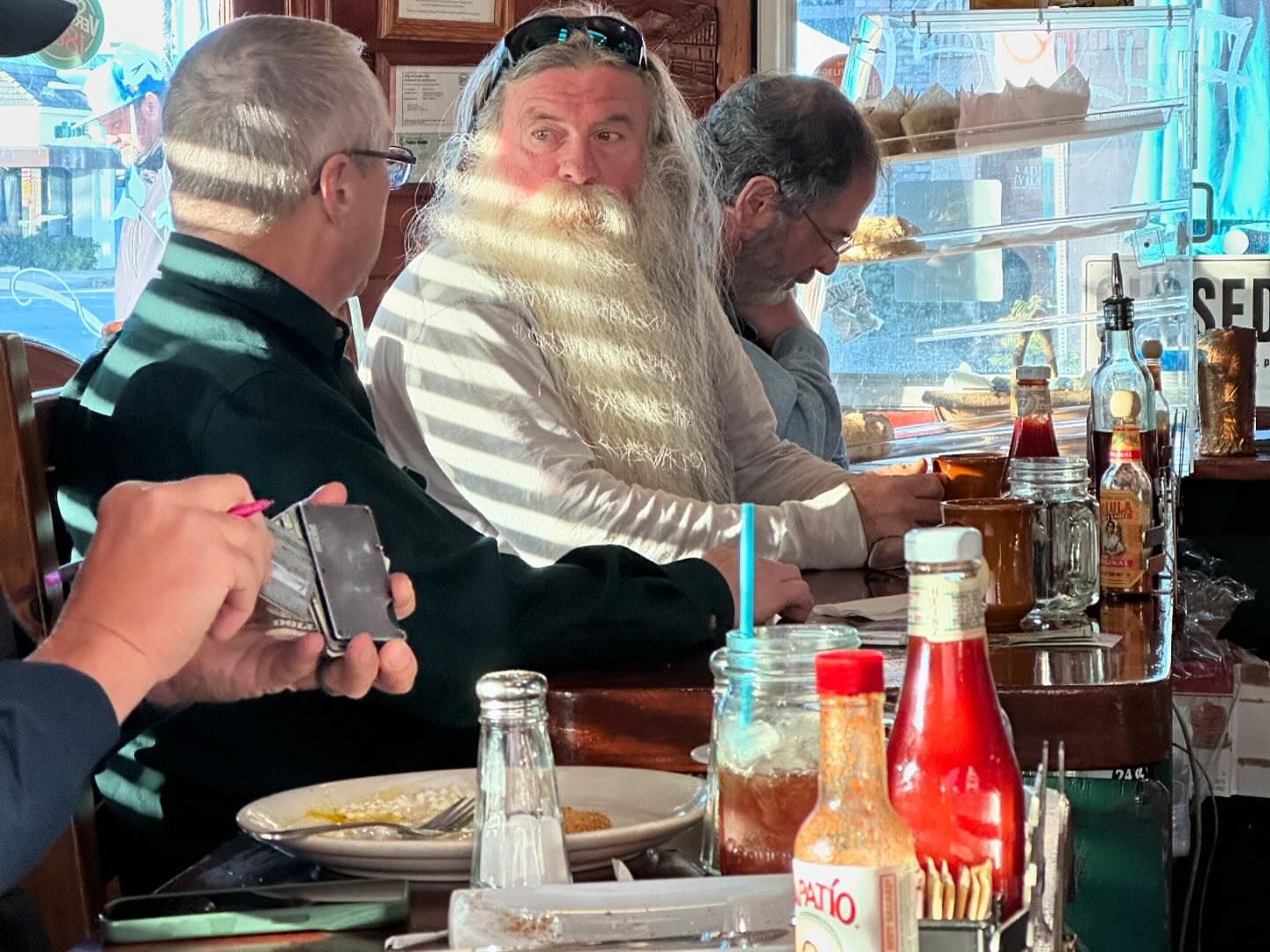 People sit at a restaurant counter with food and beverages. A man in the middle has a long white beard. Other patrons in background.