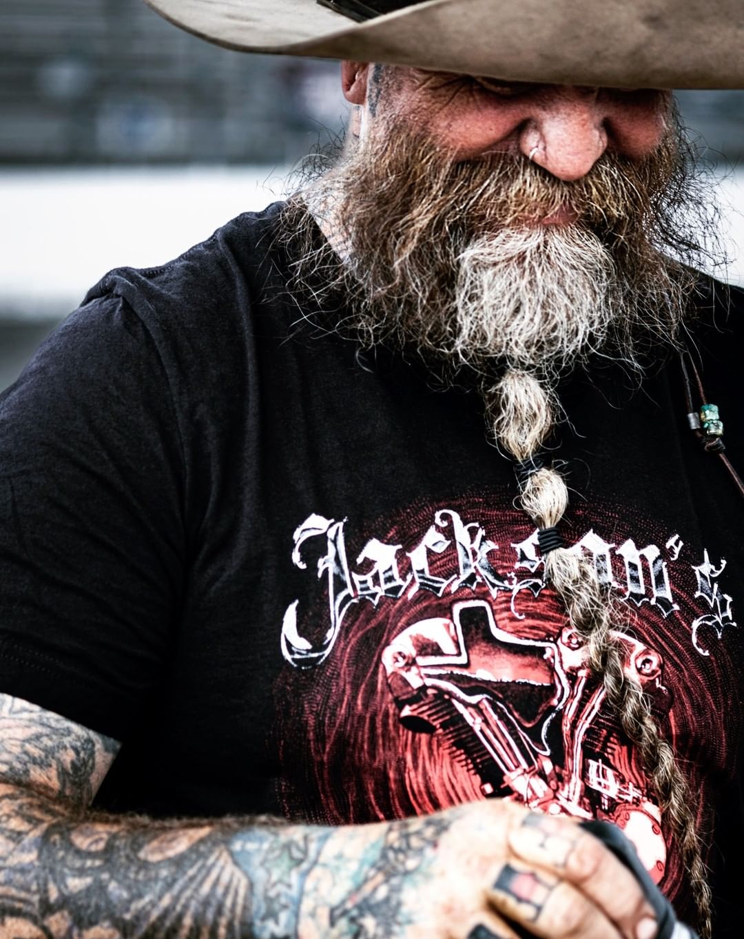 Portrait of a bearded adult male wearing a cowboy hat and t-shirt with a graphic, showcasing tattoos on his arm. The photo is tightly framed.