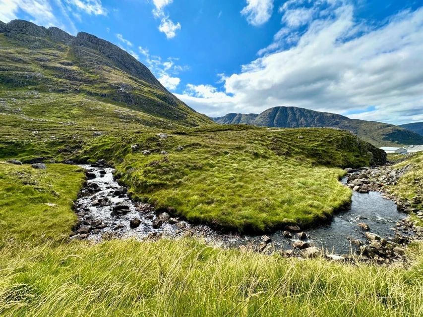 Scenic landscape featuring a stream winding through green grassy hills under a blue sky with scattered clouds, capturing the beauty of nature.