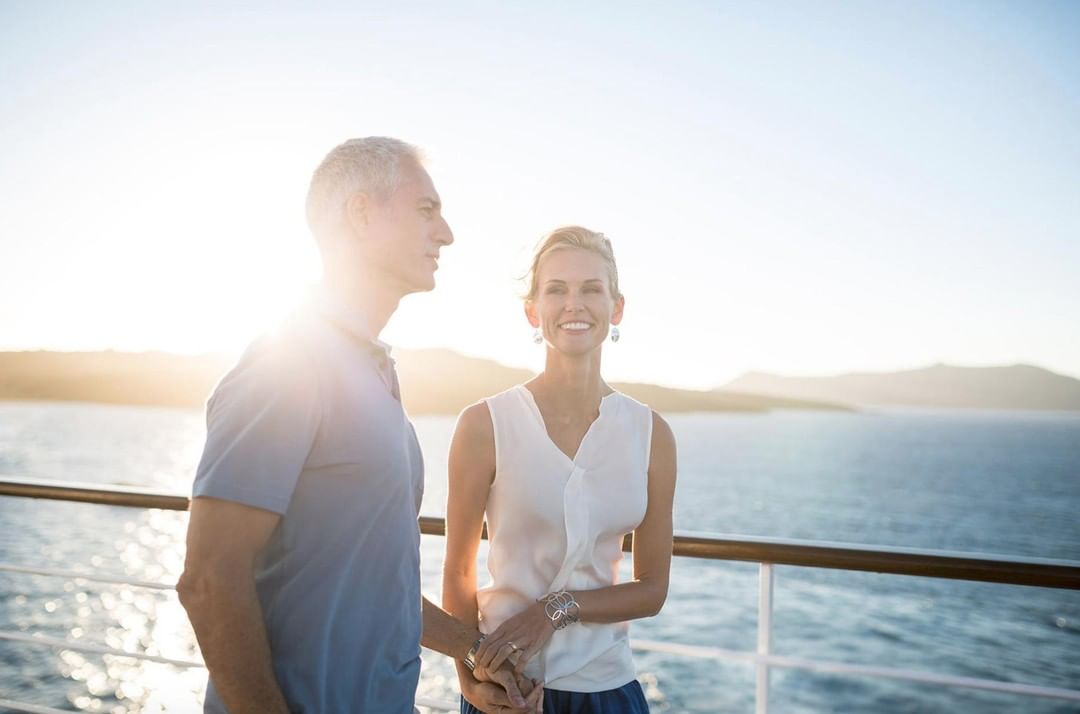 A mature couple stands on a cruise ship deck at sunset, smiling with the ocean and distant mountains in the background, creating a serene travel scene.