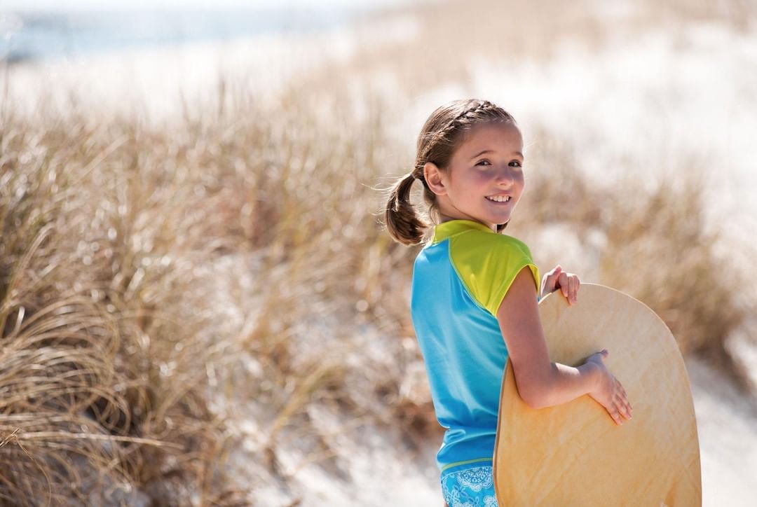 Girl with surfboard smiling at camera on beach. Child in swimwear smiles while holding board with dune grass background.