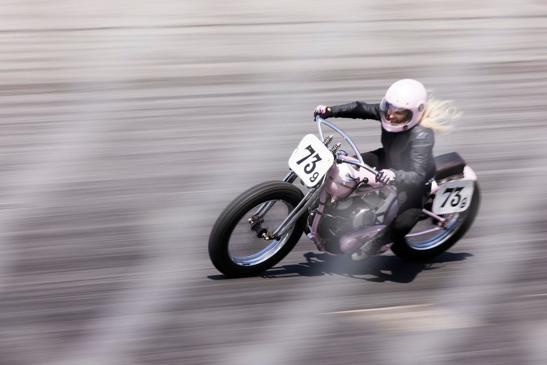 A woman races a vintage motorcycle on a track, captured in a motion blur. She wears a pink helmet and a dark suit, racing fast.