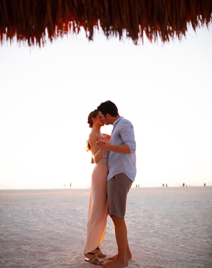 A couple shares a kiss on a sunny beach under a thatched roof, with the woman in a pink dress and the man in a blue shirt and grey shorts.