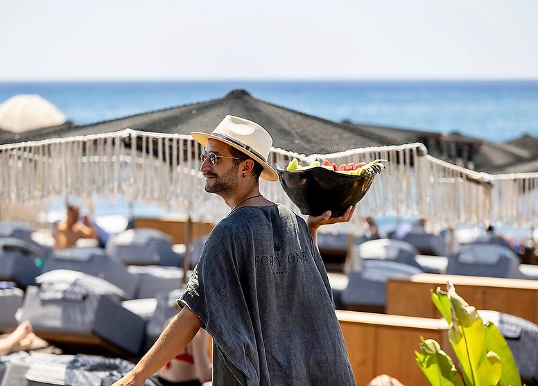 Man with a straw hat carries a bowl of fresh fruit on a sunny beach with beach umbrellas and lounge chairs near the ocean.