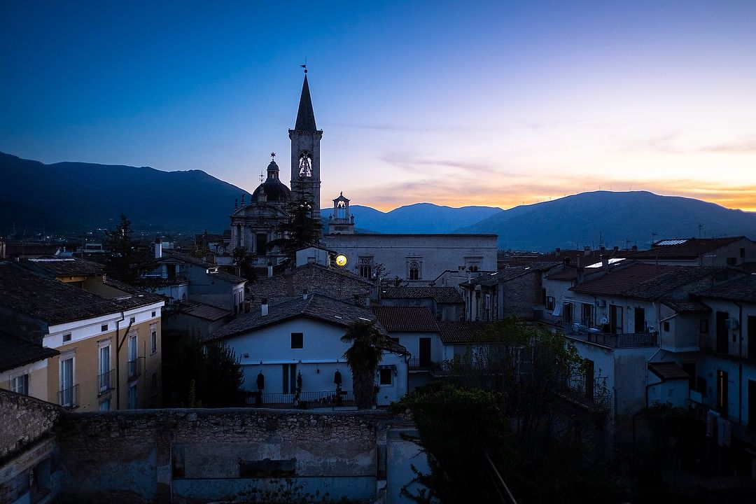 Twilight cityscape featuring a church with a bell tower, set against a backdrop of rolling mountains under a dusky sky.