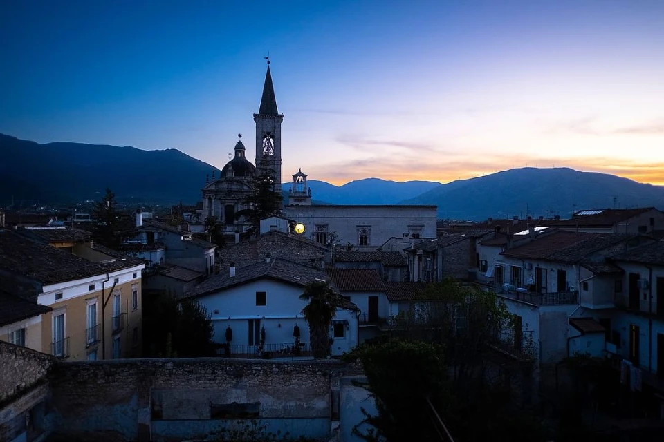 Twilight cityscape featuring a church with a bell tower, set against a backdrop of rolling mountains under a dusky sky.