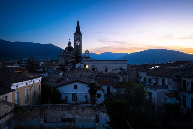 Twilight cityscape featuring a church with a bell tower, set against a backdrop of rolling mountains under a dusky sky.