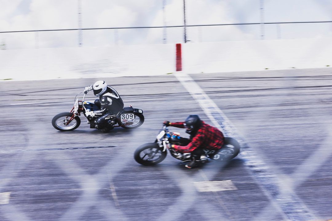 Two motorcycle racers speeding on a track, one wearing red flannel, showcasing speed and competition in an outdoor setting.