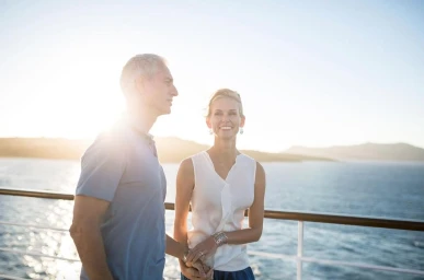 An older couple standing on a cruise ship deck at sunset, holding hands and looking out over the water.