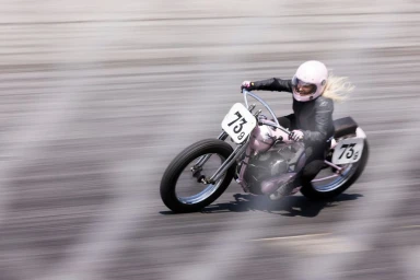A blonde woman wearing a pink helmet and black leather jacket racing a custom pink vintage motorcycle.