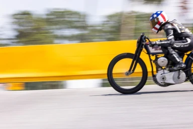 Motorcycle racer wearing an American flag helmet and black leather racing suit speeding past a yellow barrier