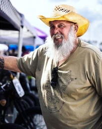 Elderly man with white beard wearing straw cowboy hat and olive t-shirt standing outdoors near motorcycles