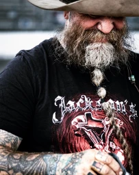 Close-up portrait of a bearded man wearing a cowboy hat, black t-shirt with graphic print, and heavily tattooed arms.