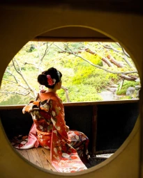 Woman in an ornate red and patterned kimono seated and looking out through a circular window framing a garden