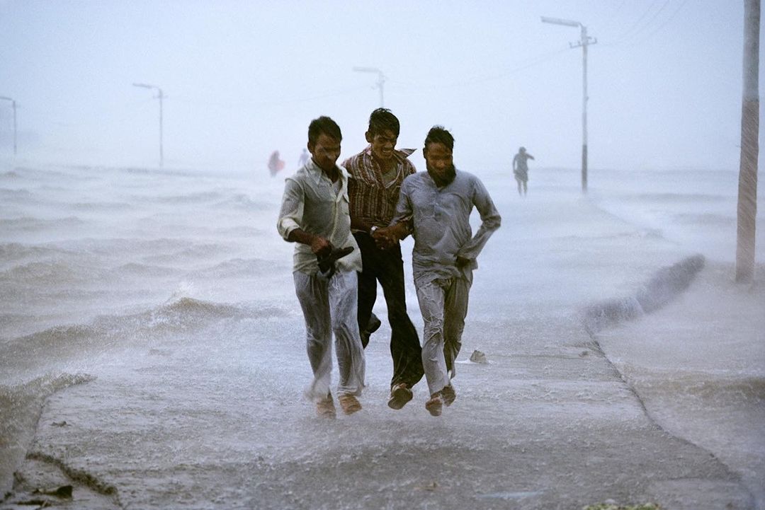Three men wade through a flooded urban street during daytime, with water reaching knee-level, power lines visible, and overcast sky.