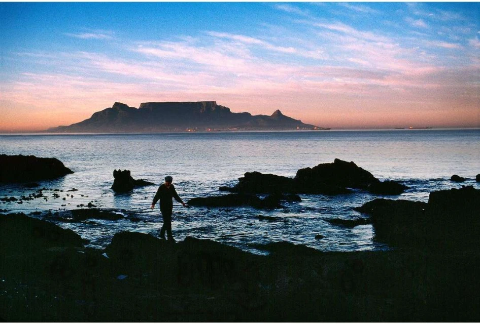 Person walking on rocks by the ocean with a mountain in the background during a colorful twilight. Coastal landscape.
