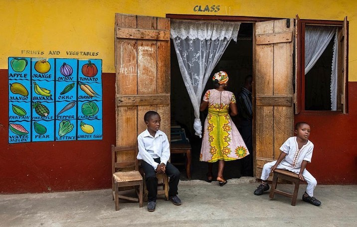 Two young African boys seated outside a classroom with a colorful educational fruits and vegetables poster on the yellow wall.