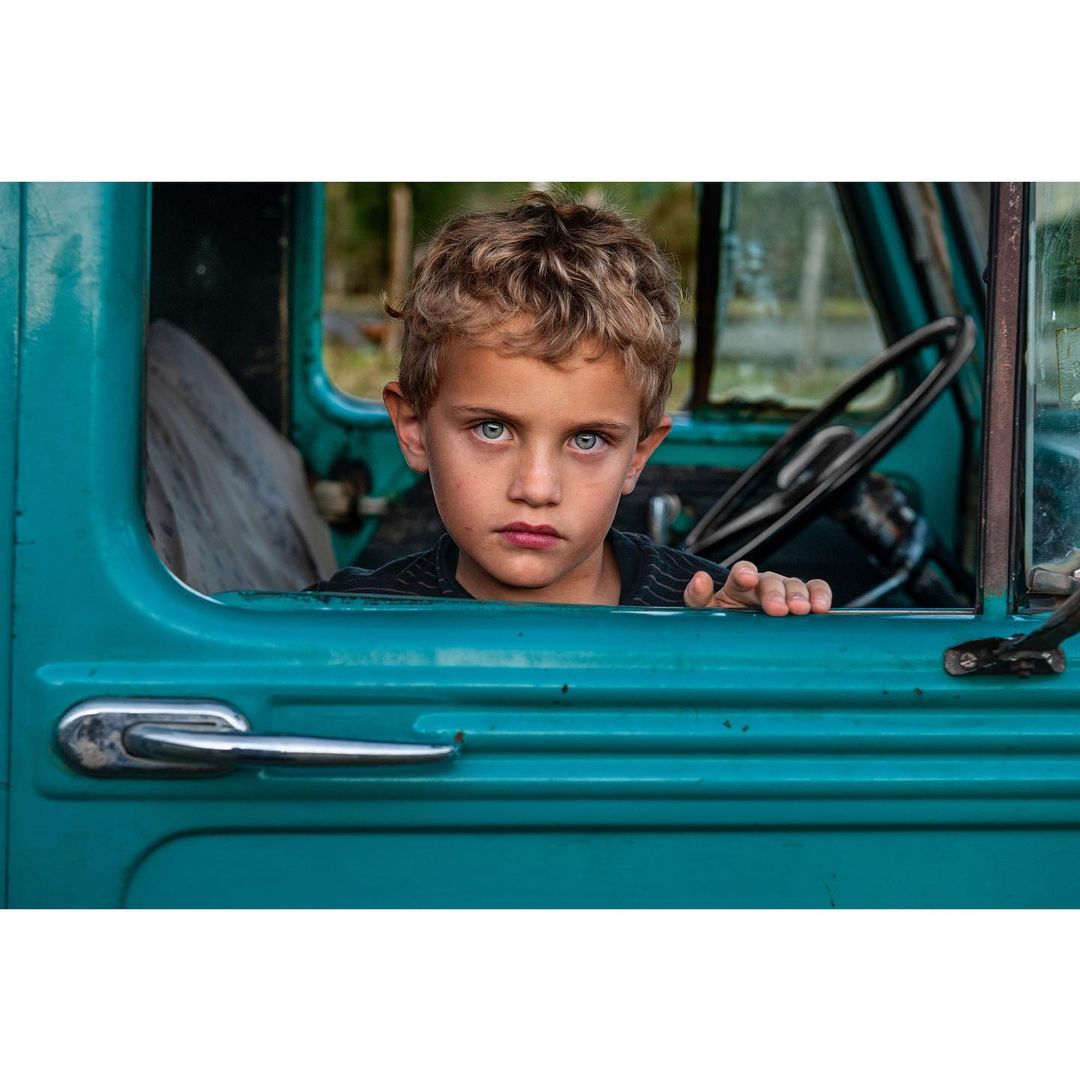 Portrait of a young boy with blond hair in a turquoise truck, looking at the camera with a serious expression, eye-level angle.
