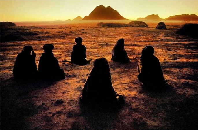 Seven silhouetted people sit in a circle on dry land with mountains visible at sunset in the background, creating a mysterious scene.