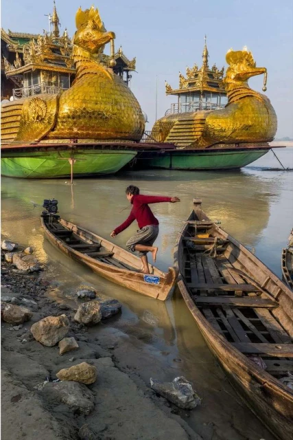 Young person leaps between small wooden boats near the riverbank; golden duck-shaped barges in the background on the river.