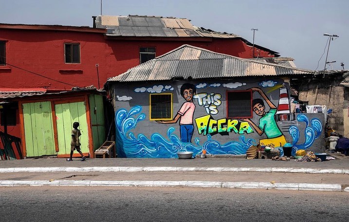 Illustration on a building with two figures and the text 'THIS IS ACCRA,' with a child walking on the sidewalk in an outdoor, urban setting.