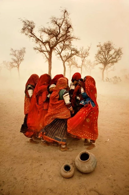 Group of women in red dresses standing in a circle in a rural landscape with bare trees and earthenware pots in the foreground.