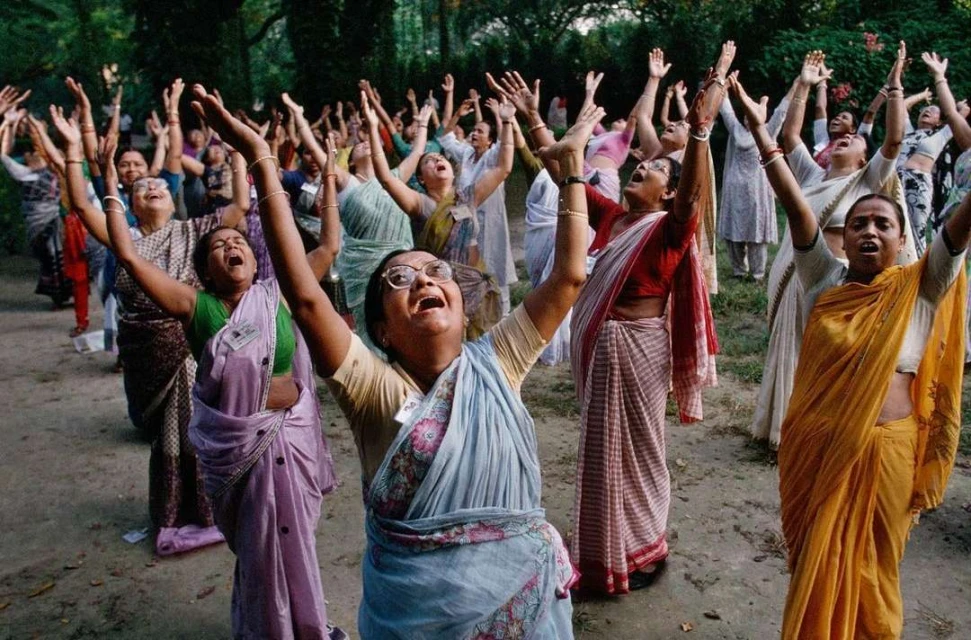 Large group of adult females outdoors with arms raised, wearing sarees, joyful expressions, looking up, greenery background.