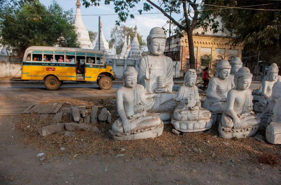 Various seated Buddha statues arranged in outdoor setting with a yellow bus and white pagodas in the background on a sunny day.