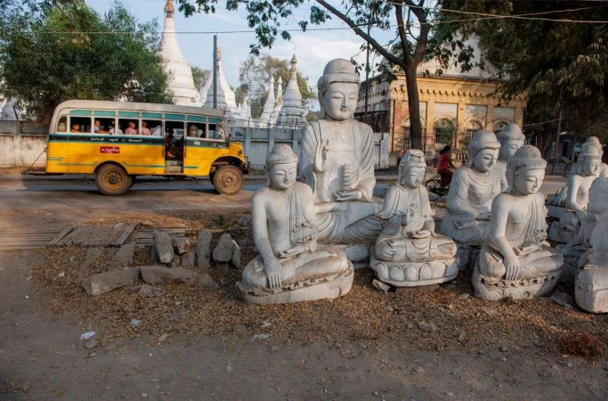 Various seated Buddha statues arranged in outdoor setting with a yellow bus and white pagodas in the background on a sunny day.