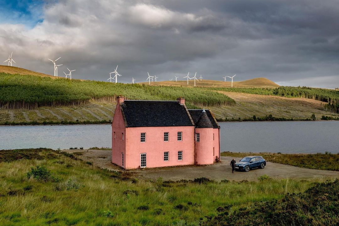 A pink house sits by a loch in Scotland with a person standing by a car. Wind turbines are on a hill in the background under a cloudy sky.