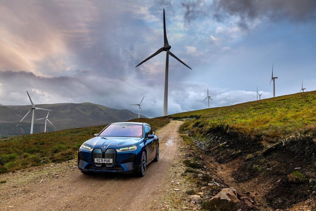 Blue SUV drives on a dirt road leading up a grassy hill, wind turbines stand in the background under a cloudy sky.